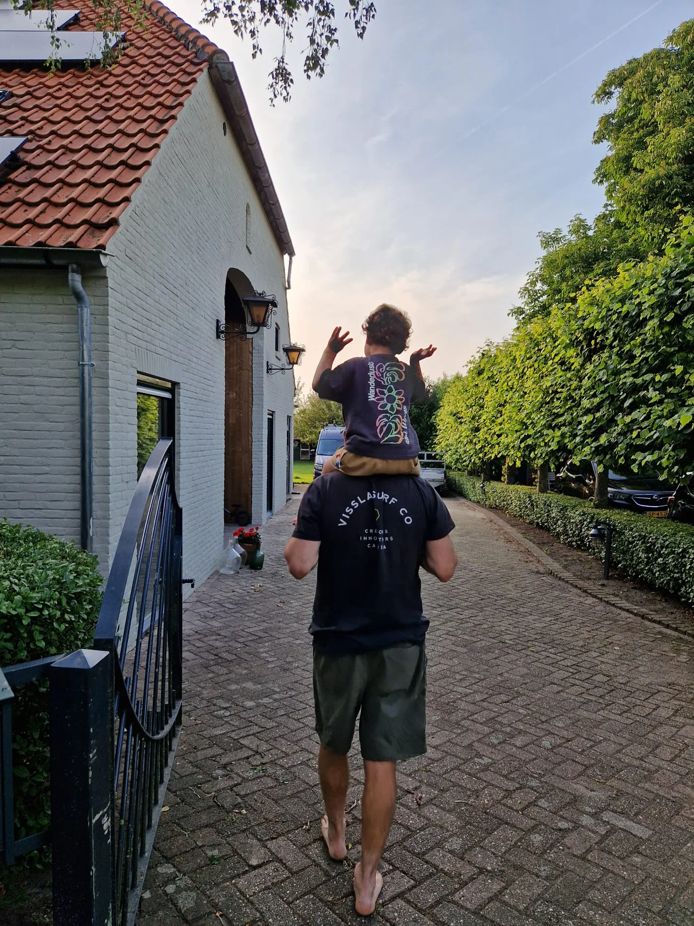 Een volwassene draagt een kind op de schouders bij een wit huis