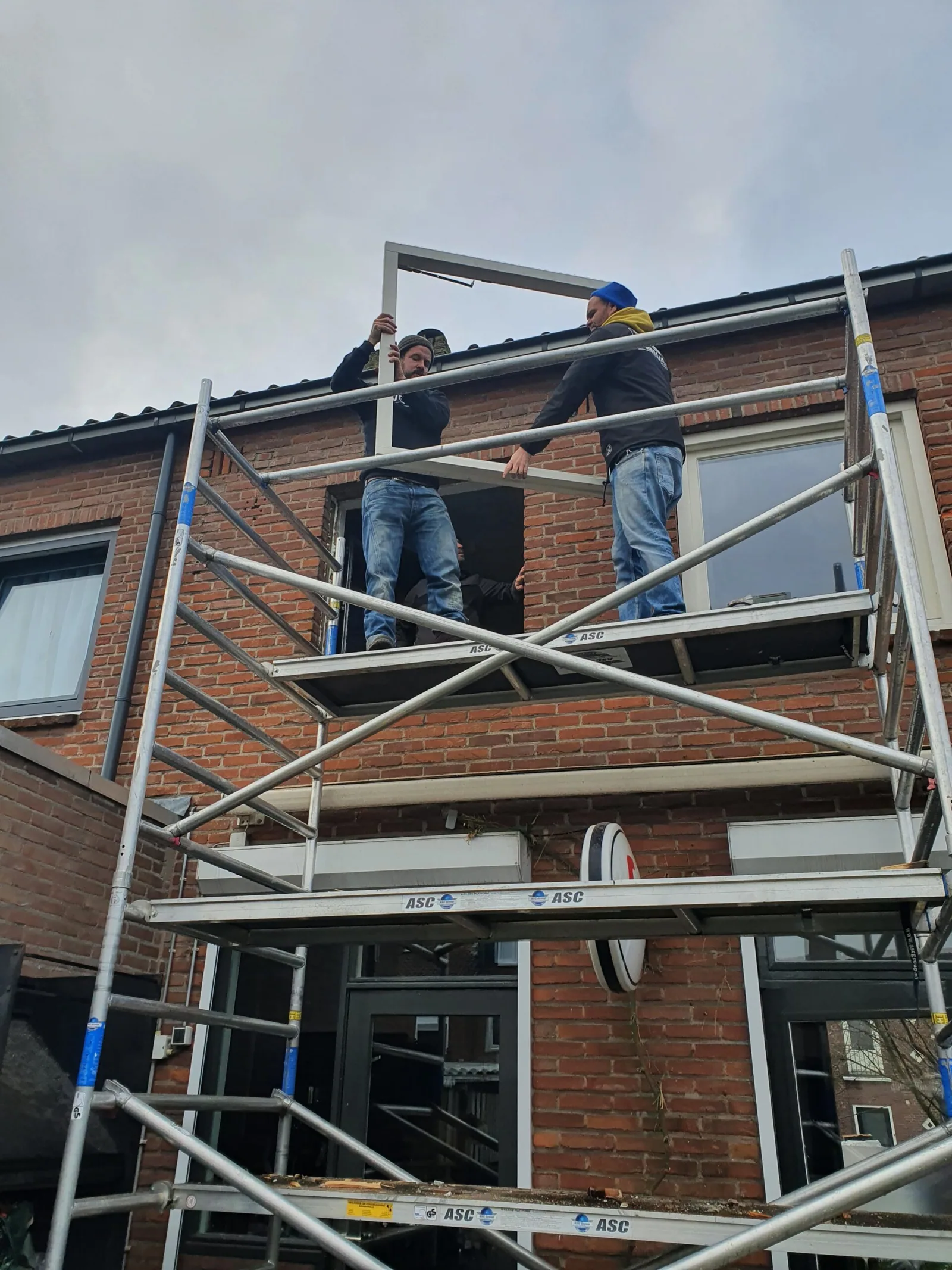 Vakmensen plaatsen een kozijn vanaf een steiger aan een bakstenen woning